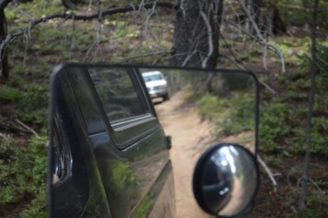 1983 Ford Bronco - Reflection in the side mirror reveals a white SUV following on a dirt path through a wooded area.