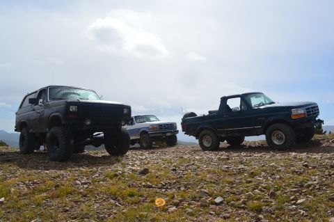 1983 Ford Bronco - Three off-road vehicles are parked on a rocky hill with a cloudy sky above.