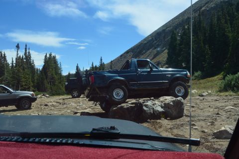 1983 Ford Bronco - Off-road vehicles navigate rocky terrain with a mountain and trees in the background.