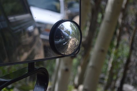 1983 Ford Bronco - Reflection in side mirror shows another vehicle on a dirt road with trees.
