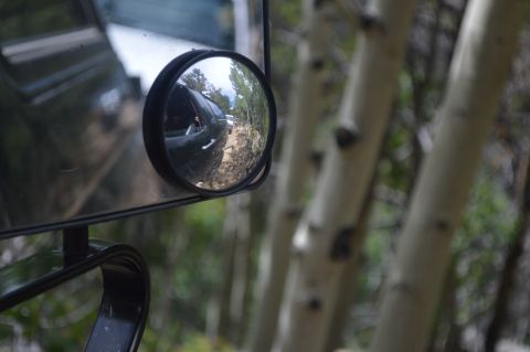 1983 Ford Bronco - A reflection in the side mirror shows a vehicle driving on a dirt road through a wooded area.