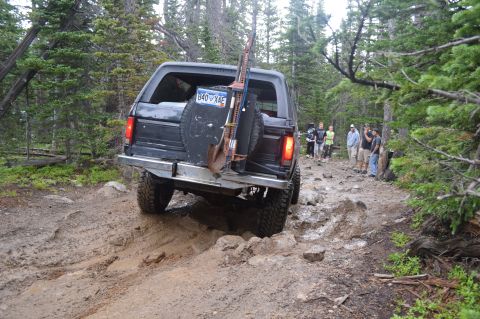 1983 Ford Bronco - A black 1983 Ford Bronco navigates a challenging rocky and muddy off-road trail, equipped with recovery tools on its rear.