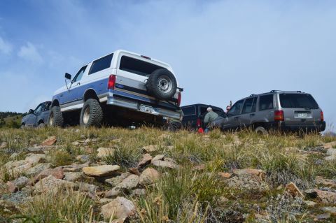 1983 Ford Bronco - Two SUVs, a Ford Bronco and a Jeep Grand Cherokee, are parked on a rocky, grassy hillside under a blue sky.