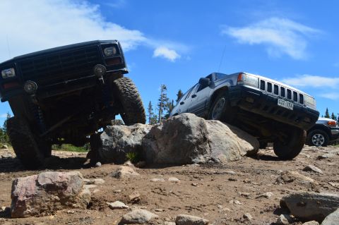 1983 Ford Bronco - Two SUVs are shown off-roading on a sunny day, navigating a challenging rocky course.