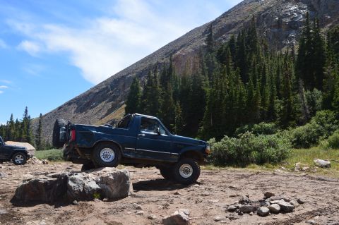 1983 Ford Bronco - A dark blue SUV navigates a rocky mountain trail, with a spare tire mounted on the rear. Another vehicle is visible to the left.