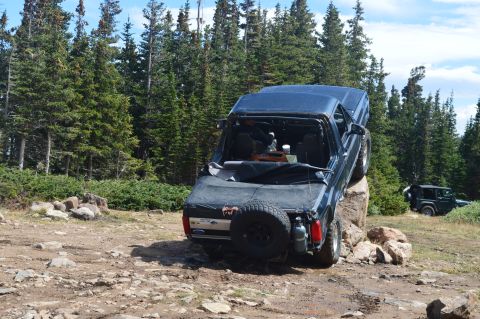 1983 Ford Bronco - A dark blue SUV is precariously balanced on a large rock on a dirt trail, with a forest in the background.