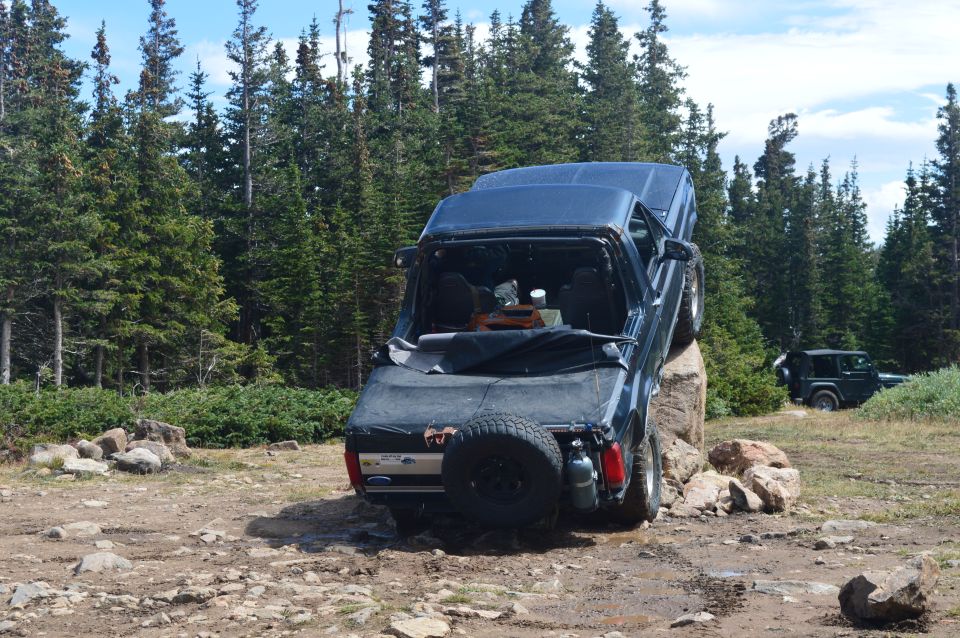 1983 Ford Bronco with spare tire on rear, positioned on a large rock off-road, another Jeep visible in background