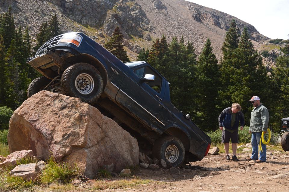 1983 Ford Bronco climbing a large rock on a mountain trail with two men watching