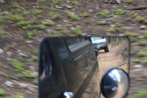 1983 Ford Bronco - A silver SUV is seen driving on a dirt road, reflected in the side mirror of another vehicle.