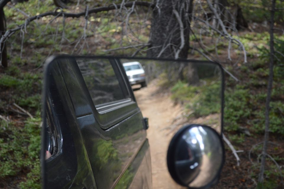 Reflection in side mirror shows a white SUV driving on a dirt road behind the camera vehicle