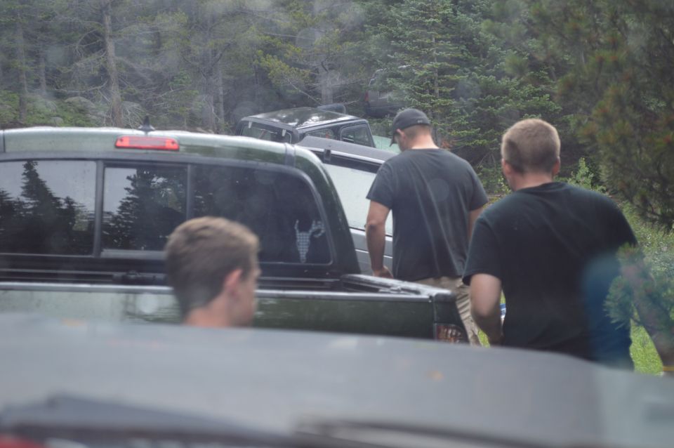 Several people stand around pickup trucks and SUVs parked in a wooded area