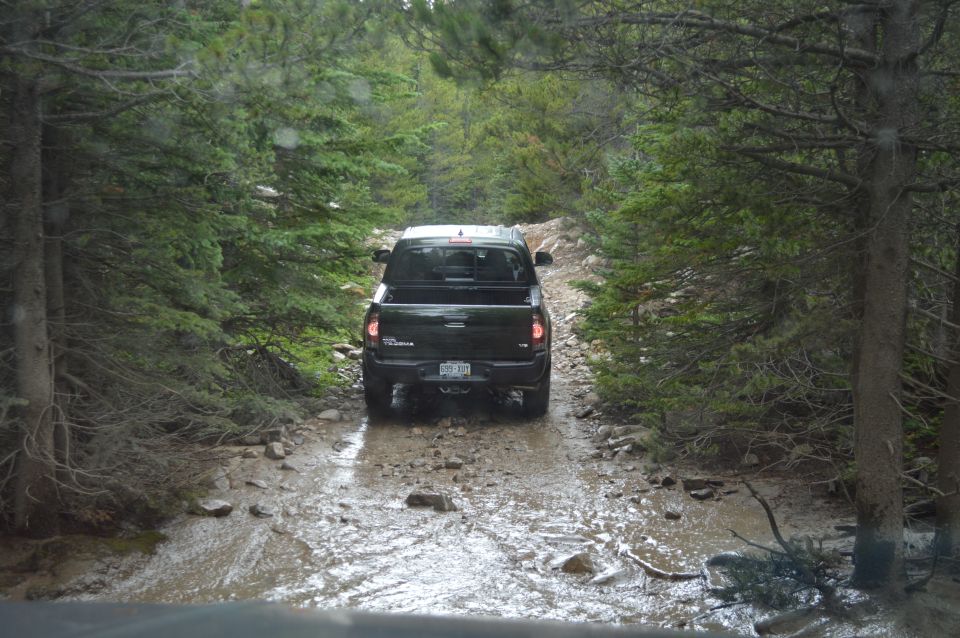 Black pickup truck driving on a muddy, rocky forest road with trees on both sides