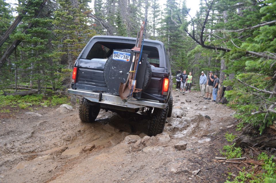Black 1983 Ford Bronco driving on a rocky, muddy trail with a shovel and jack mounted on the back. People watch from the side of the trail