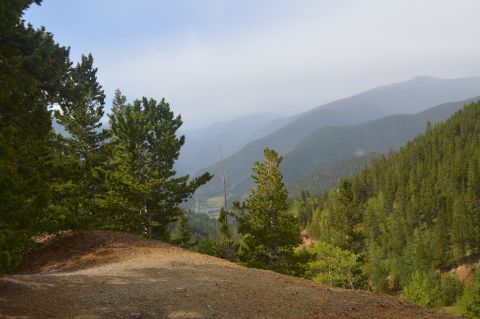 1983 Ford Bronco - A scenic view of rolling mountains and dense forests, with a dirt path in the foreground and a Supermotors logo at the bottom right.