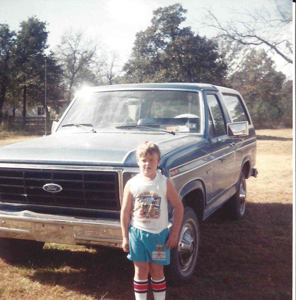 Boy in shorts and striped socks stands before a blue 1980 Ford Bronco