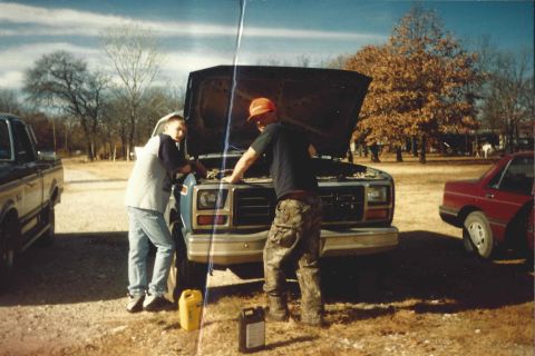1982 Ford Bronco - Two men are working on the engine of a blue truck outdoors, with other vehicles visible in the background.