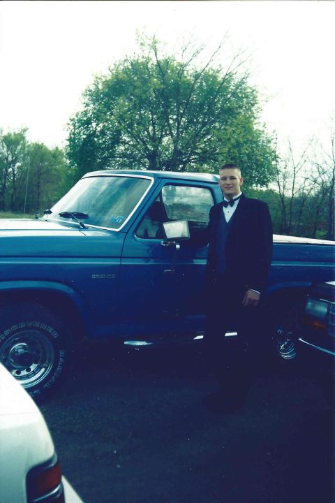 1982 Ford Bronco - A man in a tuxedo poses next to a blue Ford Bronco, with another vehicle partially visible in the foreground.