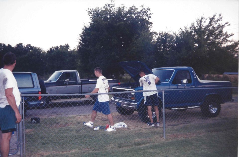 Two men are working on the engine of a blue pickup truck. A second pickup truck is parked to the left. Fence in foreground