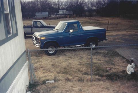 1982 Ford Bronco - A blue Ford Bronco from the early 1980s is parked in a yard with another truck visible in the background.