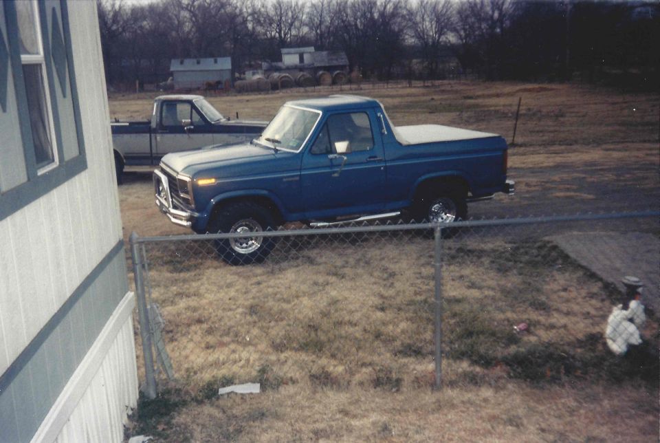 Blue 1980s Ford Bronco with a white soft top, parked in a grassy yard next to a trailer home