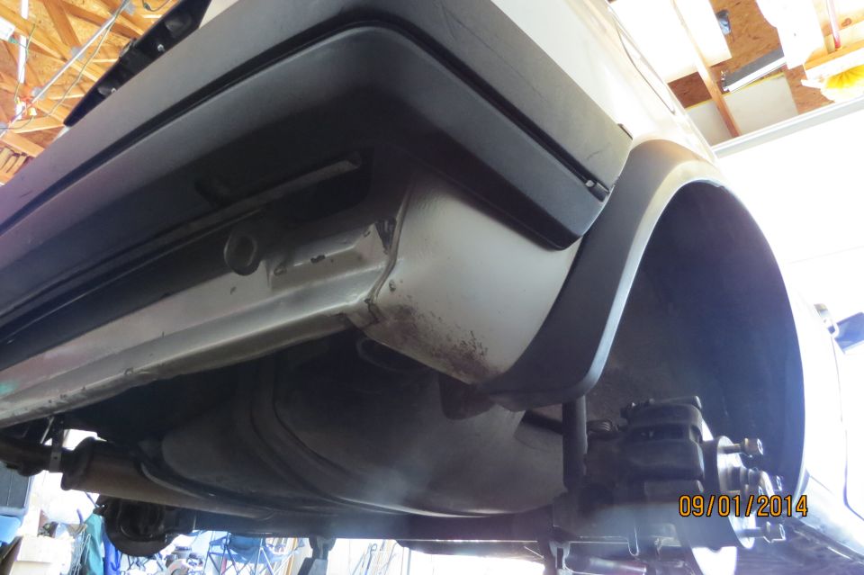 Underside view of a car's rear bumper and wheel well, showing rust and suspension components