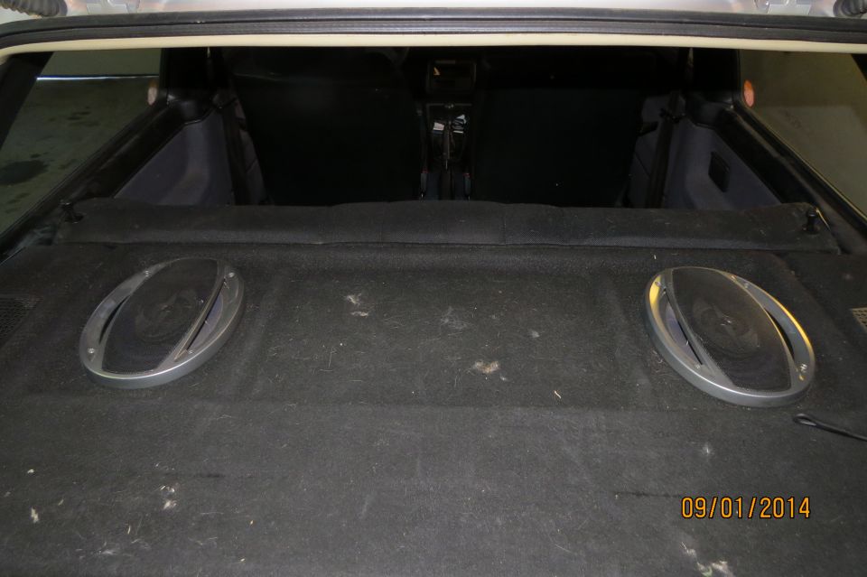 Rear cargo area of a car showing two oval speakers mounted on the carpeted floor. Interior seats are visible in the background