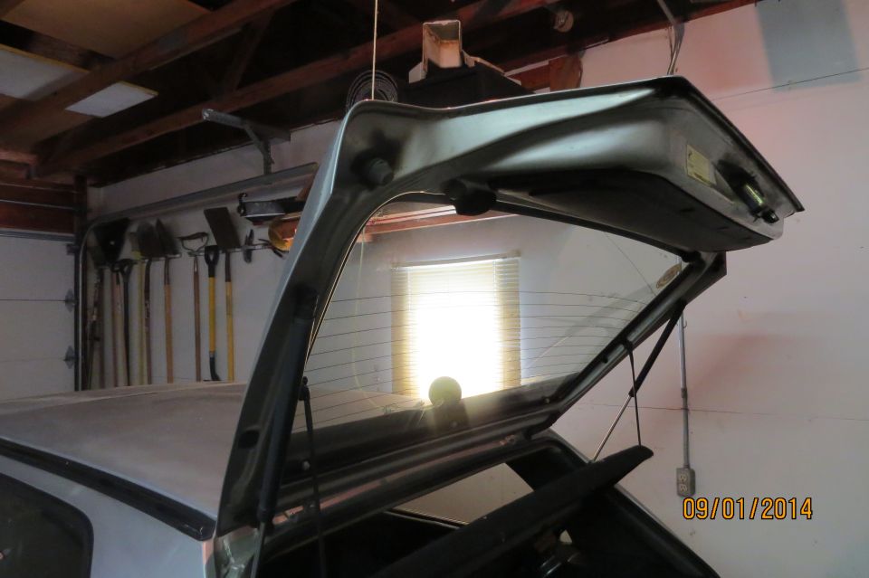 Car's rear hatch open, showing the back window and interior. Garage setting with tools on wall