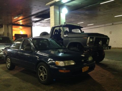 1979 Ford Bronco - A dark pickup truck with a light bar is parked behind a sedan in a garage.