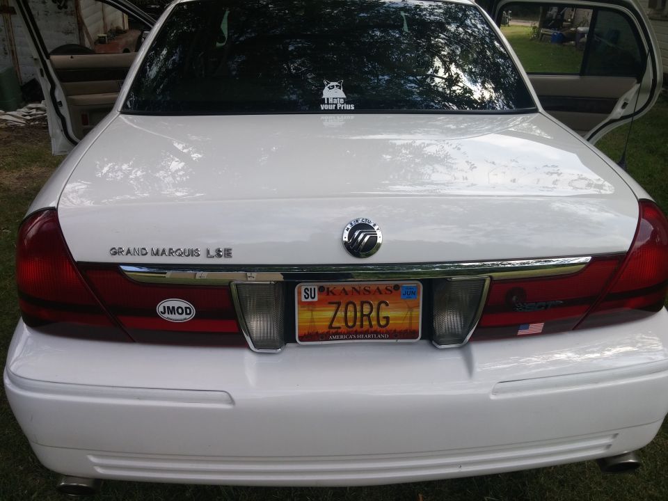 Rear of a white 2000 Mercury Grand Marquis with a Kansas license plate that reads ZORG. It has a 'JMOD' sticker and a cat sticker on the rear window