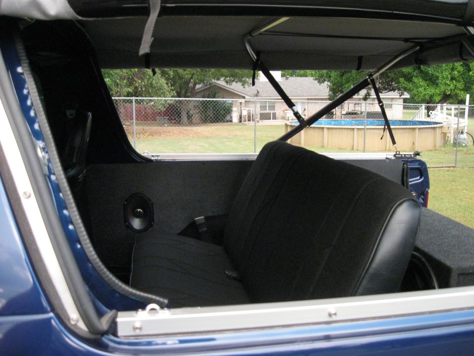 Interior view of a blue SUV showing the rear seat, speakers, and a subwoofer box. Backyard with a pool is visible through the window