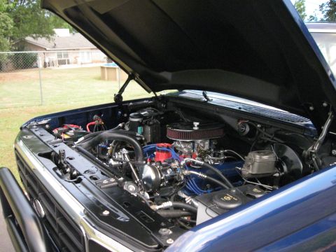 1982 Ford Bronco - The engine bay of a blue 1982 Ford Bronco is shown with the hood open, revealing a clean engine with chrome accents and blue spark plug wir…