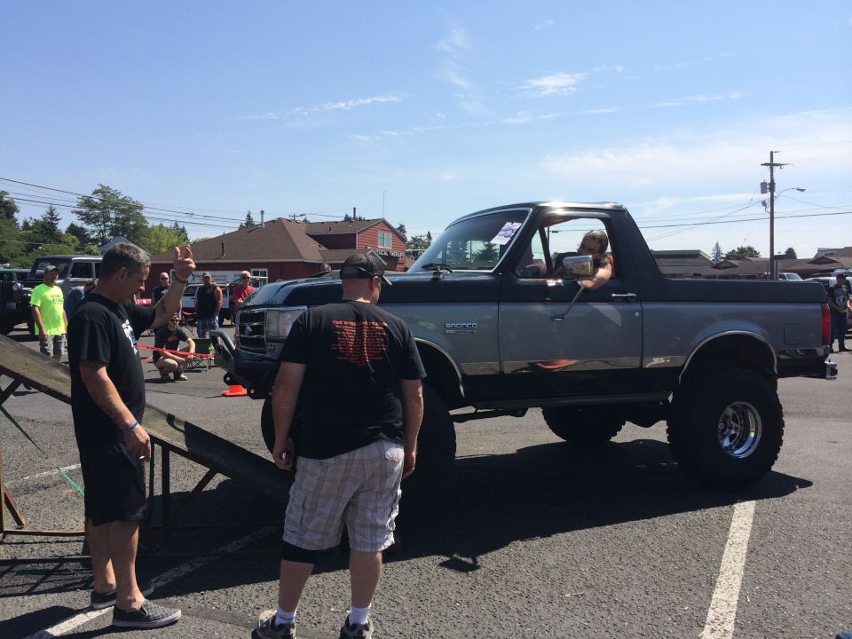 1989 Ford Bronco lifted with large tires and custom paint, parked outdoors with people around