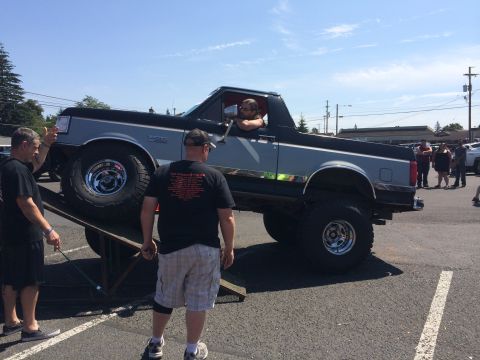 1989 Ford Bronco - A lifted pickup truck is positioned on ramps at an outdoor event with several people observing.