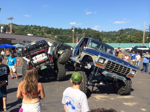 1989 Ford Bronco - A lifted blue and silver Ford Bronco, likely from the late 1970s, is displayed at an off-road show with another modified vehicle.