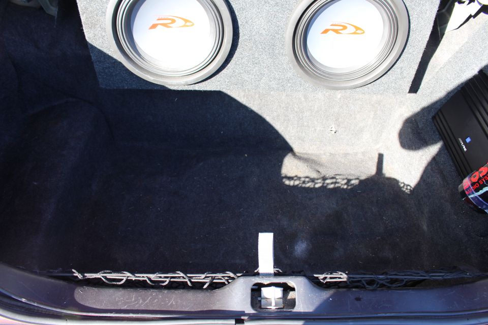 Car trunk interior with two subwoofers and an amplifier, dark carpet lining, and a cargo net