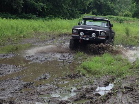1979 Ford Bronco - A black 1979 Ford Bronco navigates a muddy off-road trail, kicking up water and dirt.