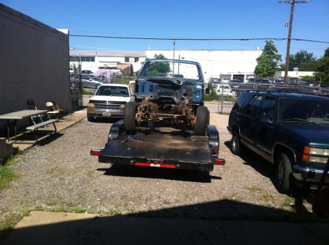 1982 Ford Bronco - A blue truck chassis is loaded onto a trailer, with a white pickup and a dark SUV parked in the vicinity.