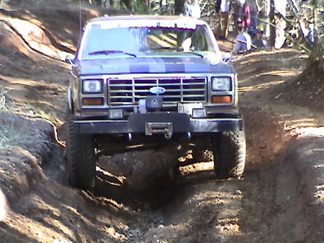 Lifted brown Ford with winch bumper and off-road tires crawling a rutted dirt trail in the woods, front view