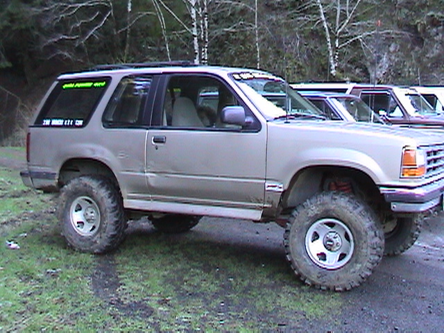 Lifted tan two-door SUV with large mud tires, side view, parked on grass with trees behind