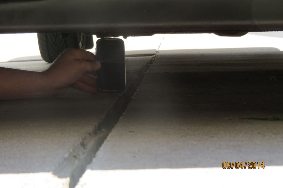 Hand holding smartphone under car chassis, near a crack in pavement