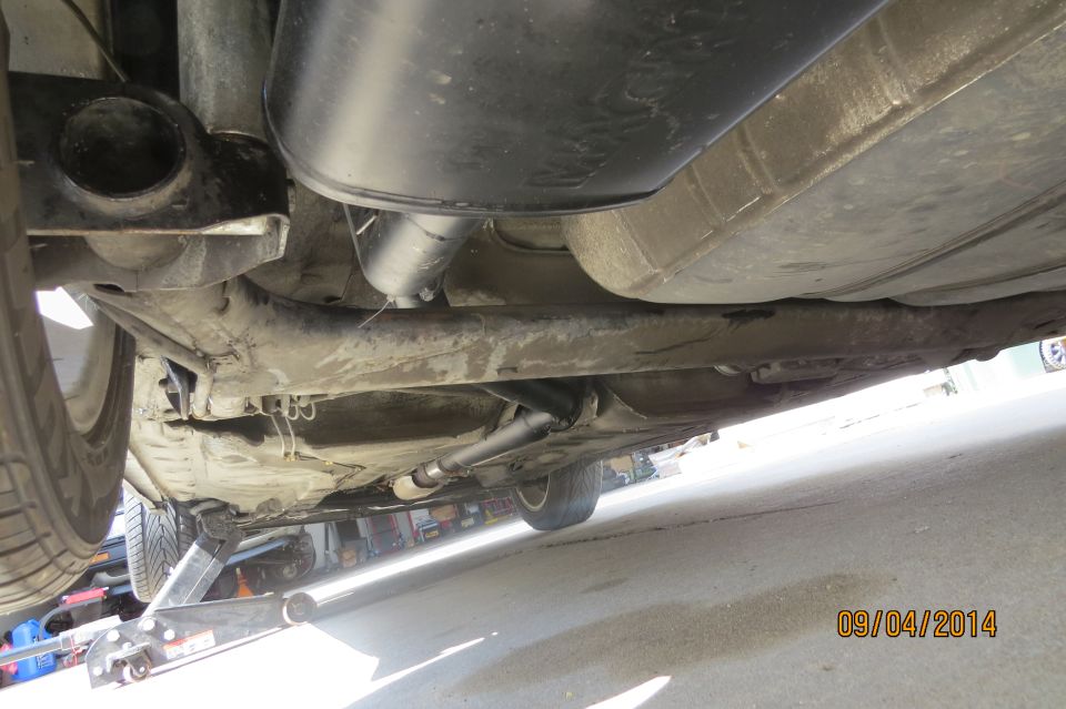 Underside view of a car showing the exhaust pipe, muffler, and fuel tank. A jack is visible