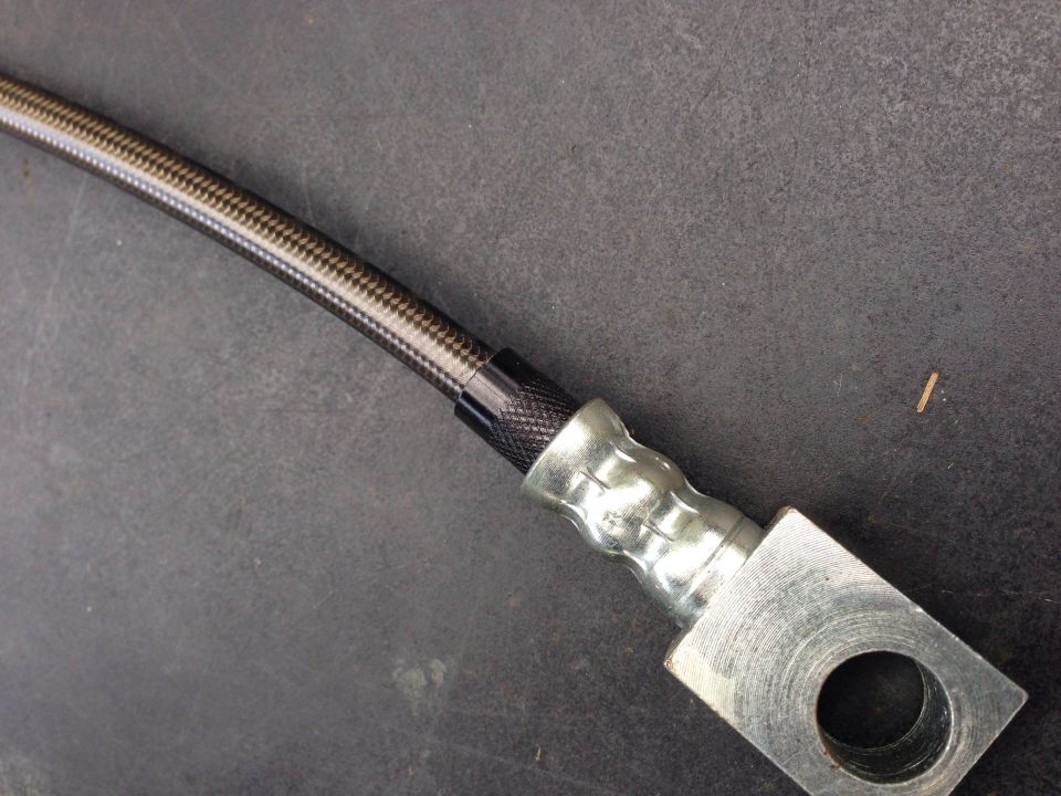 Close-up of a braided brake hose with metal fittings. The hose has a carbon fiber pattern, and the fittings are silver metal