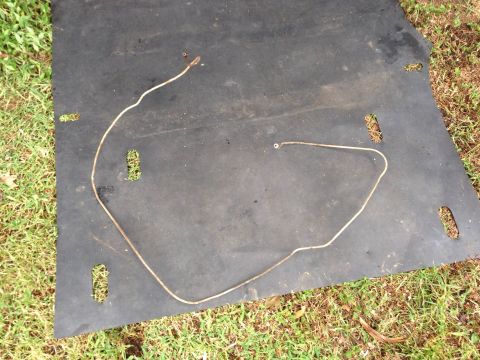 1979 Ford Bronco - A metal brake line lies on a black mat, with grass visible around the edges. The Supermotors.net logo is in the lower right corner.