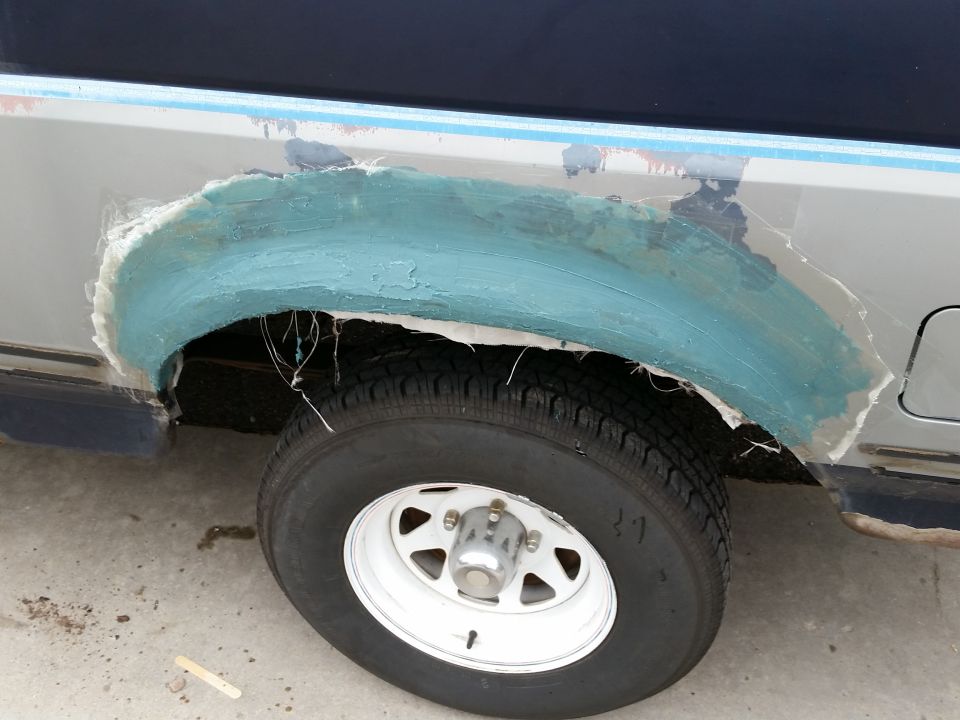 Close-up of a vehicle's rear fender area with fiberglass and blue filler being applied over a damaged section. A white-rimmed tire is visible