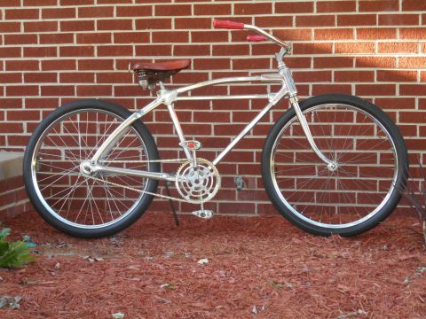Chrome cruiser bicycle with red handlebars and a brown leather seat, parked against a brick wall
