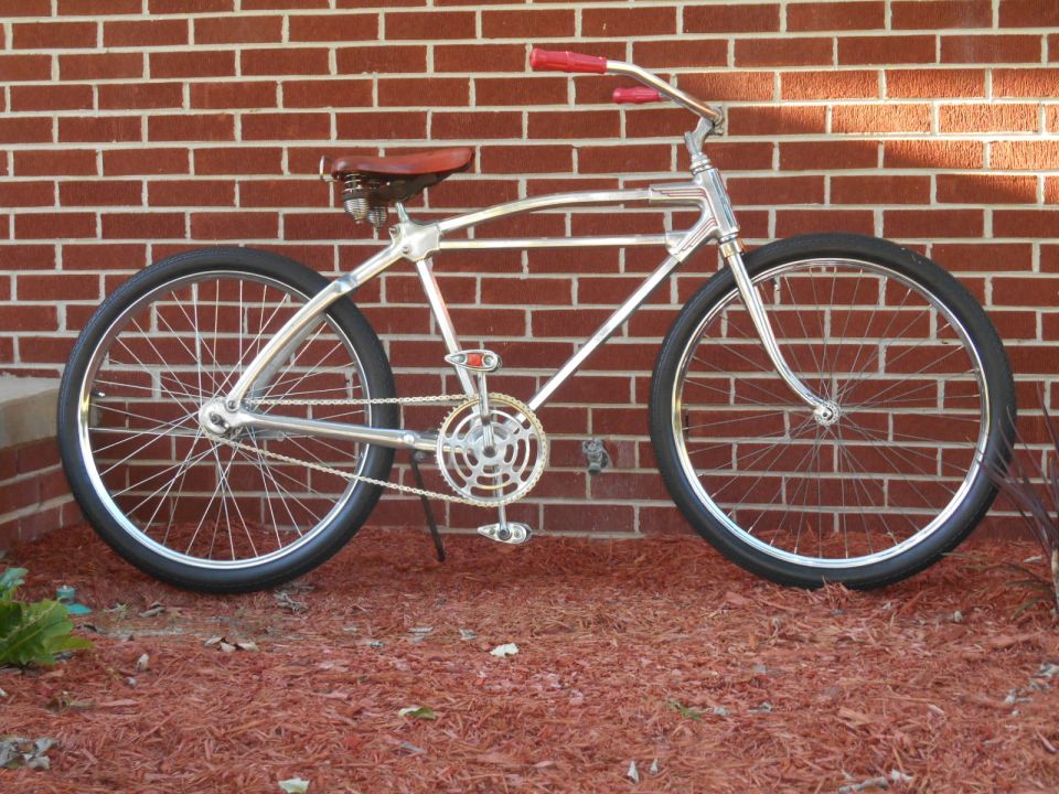 Chrome cruiser bicycle with red handlebars and a brown leather seat, parked against a brick wall