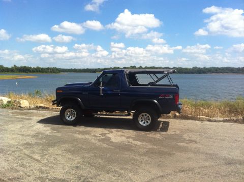 1982 Ford Bronco - A blue Ford Bronco is parked on a paved area next to a body of water under a partly cloudy sky.