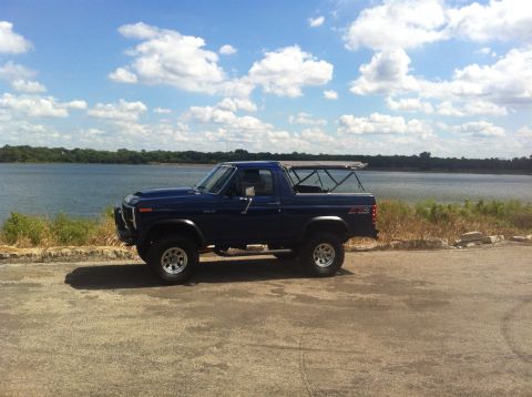 1982 Ford Bronco - A blue Ford Bronco is parked on a paved area next to a body of water under a cloudy sky.