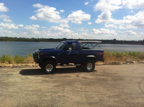 1982 Ford Bronco - Side view of a blue Ford Bronco parked near a body of water under a cloudy sky.
