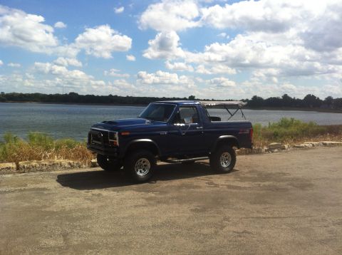 1982 Ford Bronco - A blue 1980 Ford Bronco sits parked on a paved area next to a body of water under a cloudy sky.